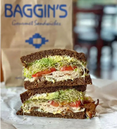 Hearty stacked rye turkey sandwich cut in half, layered with tomato, bacon and alfalfa sprouts on deli paper with a blurred café bag and interior in the background.