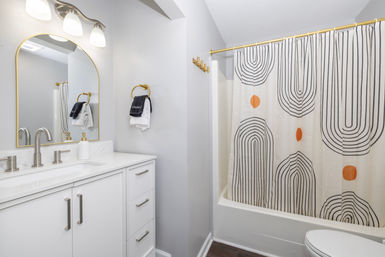Bright modern bathroom with white vanity and marble-look countertop, gold-accent arched mirror and fixtures, patterned black-and-orange shower curtain and light gray walls.