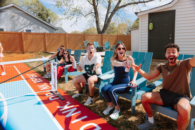 Group of friends cheering and holding drinks in blue Adirondack chairs at a sunny backyard next to a colorful red-and-blue portable net court.
