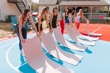 Six women in colorful activewear unrolling pale yoga mats on a sunny blue-and-red outdoor basketball court beside suburban houses, smiling as they prepare for a group workout.