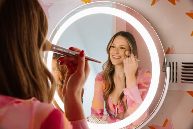 Smiling woman applying blush with a makeup brush in front of an illuminated round vanity mirror and ring light, wearing a pink patterned top against a star-accented wall — playful beauty routine.