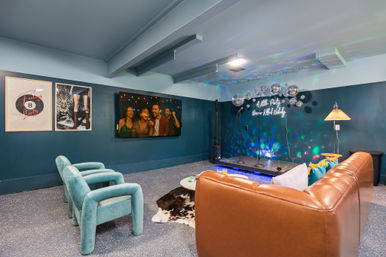Cozy basement karaoke lounge with a small raised stage, cluster of disco balls and neon sign, leather sectional, teal velvet armchairs, wall-mounted TV and colorful party lights.