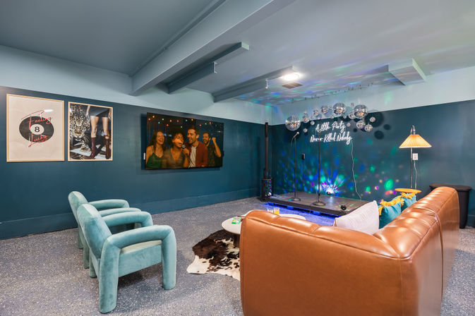 Cozy basement karaoke lounge with a small raised stage, cluster of disco balls and neon sign, leather sectional, teal velvet armchairs, wall-mounted TV and colorful party lights.