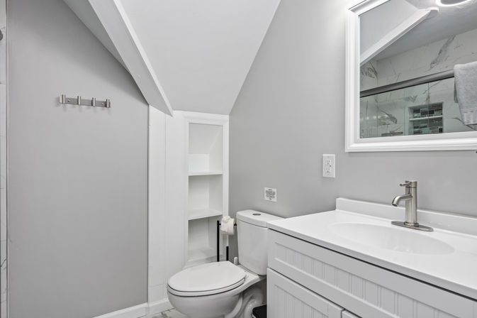 Cozy modern gray-and-white small attic bathroom with sloped ceiling, white vanity and sink with chrome faucet, toilet next to built-in shelving, and mirror reflecting a marble-tiled shower.