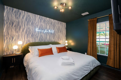 Modern guest bedroom with king bed, green velvet headboard, white linens and orange pillows, patterned accent wall with neon sign, round bedside lamps, mustard curtains and folded towels on bed.