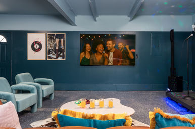 Cozy indoor karaoke lounge interior with teal armchairs, wall-mounted TV showing friends singing, small stage with microphone, coffee table with drinks and colorful cushions