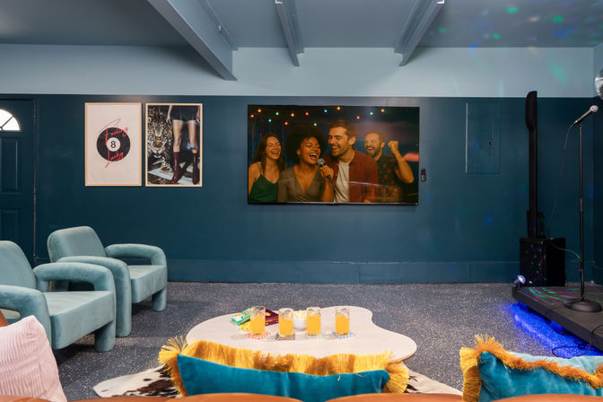 Cozy indoor karaoke lounge interior with teal armchairs, wall-mounted TV showing friends singing, small stage with microphone, coffee table with drinks and colorful cushions