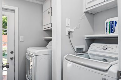 Bright compact laundry room with white top‑load washer and matching dryer, built‑in shelves and cabinet, detergent on shelf, and a glass back door showing a sunny yard.