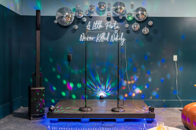Indoor party stage with two microphone stands, mirrored disco balls and colorful spotlights on a teal wall, neon sign reading 'A Little Party Never Killed Nobody', tall speaker and floor uplight.