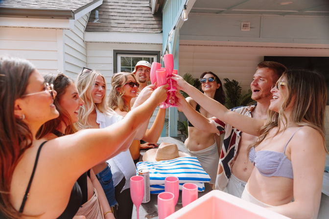 Sunny backyard poolside patio party with friends in swimsuits and sunglasses raising pink plastic champagne flutes in a cheerful toast, striped beach towel and drinks on the counter