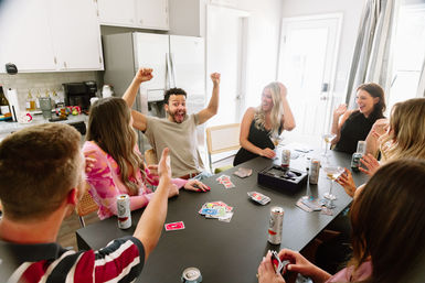 Group of friends laughing around a black dining table in a bright modern kitchen, celebrating while playing a colorful card game with cocktails and canned drinks