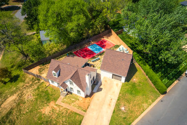 Aerial shot of a suburban home and detached garage with a fenced backyard featuring a colorful red-and-blue half-court basketball court, patio seating, concrete driveway and surrounding green trees