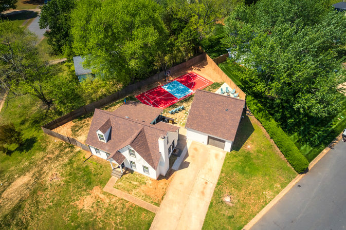 Aerial shot of a suburban home and detached garage with a fenced backyard featuring a colorful red-and-blue half-court basketball court, patio seating, concrete driveway and surrounding green trees