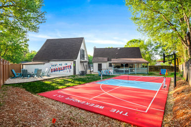 Sunny Charlotte backyard with a bright red multipurpose court (pickleball/basketball) with hoop and net, two white homes with covered porch and 'Meet Me in Charlotte' mural, Adirondack seating area and leafy trees