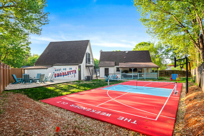 Sunny Charlotte backyard with a bright red multipurpose court (pickleball/basketball) with hoop and net, two white homes with covered porch and 'Meet Me in Charlotte' mural, Adirondack seating area and leafy trees