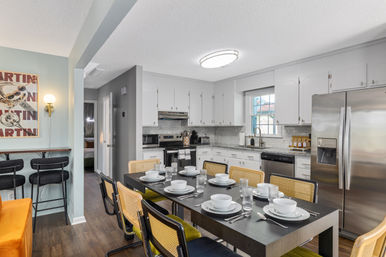 Open-concept modern kitchen and dining area — black table set for eight with white dishware, cane-back chairs with green cushions, white cabinets, subway-tile backsplash, stainless-steel appliances and a small breakfast bar