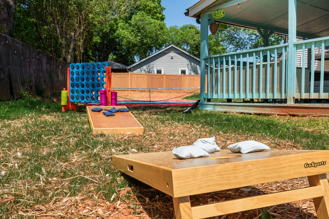 Sunny suburban backyard lawn games: wooden cornhole boards with bean bags and a giant Connect Four next to a teal covered porch.