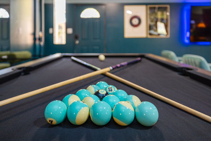 Teal-and-pale-yellow striped billiard balls neatly racked on a black-felt pool table with crossed cues in a cozy indoor game room.
