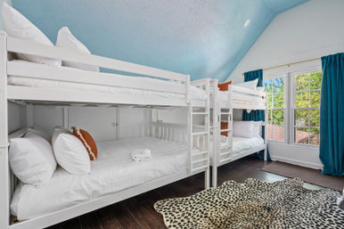 Cozy loft guest room with white double bunk beds, blue vaulted ceiling, teal curtains, dark hardwood floors, leopard-print rug and large window with leafy view.