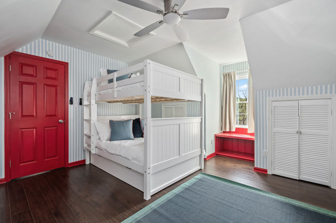 Bright attic bedroom with white wooden bunk bed, red door and window bench, pale blue striped walls, ceiling fan and dark hardwood floors