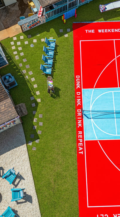 Aerial shot of a vibrant red-and-blue backyard basketball court in a residential yard, surrounded by green lawn, turquoise Adirondack chairs around a fire pit, stepping stones and a nearby house roof.