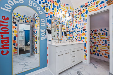 Bright modern bathroom with a white double-sink vanity, marble-look floor, gold fixtures, large arched statement mirror with a playful painted slogan, and bold multicolor abstract brushstroke wallpaper extending into an adjoining room.