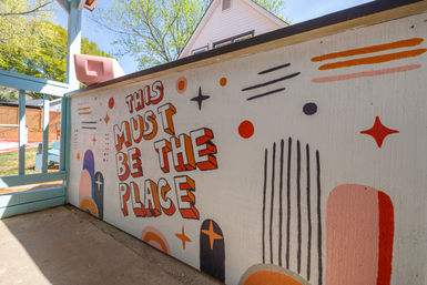 Sunny backyard patio wall mural reading "THIS MUST BE THE PLACE" in bold 3D orange lettering with playful stars, dots and pastel arches on a deck railing.