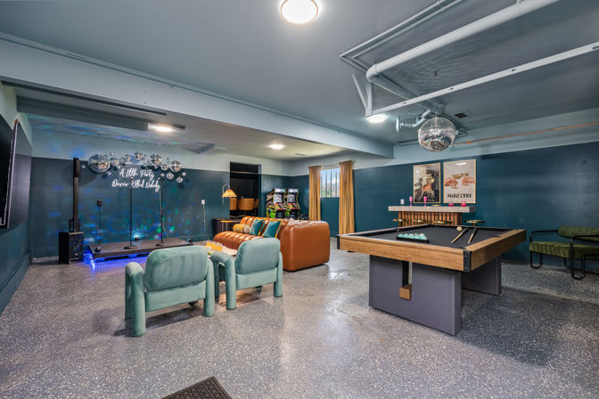 Residential basement game room with wooden pool table, disco ball, leather sofa and teal lounge chairs, small stage with neon sign reading "A little party never killed nobody", arcade machines and colorful lighting