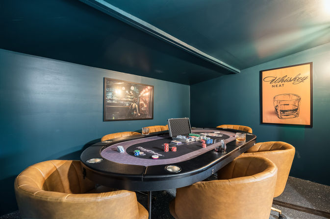 Cozy basement poker room with teal walls, tan leather swivel chairs around a felt poker table set with chips, cards and drinks, plus framed bar photo and Whiskey Neat poster.