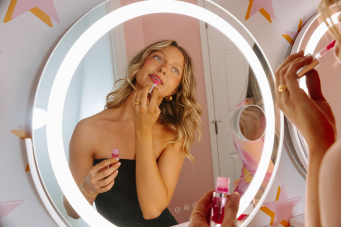 Woman applying shiny pink lip gloss at an illuminated round vanity mirror with LED ring light in a pastel pink room decorated with star decals