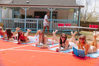 Group of women in swimsuits lounging on striped towels on a red backyard sports court, sunbathing with sunglasses and pink drinks while a man walks by a porch and a folding table set up with red beer-pong cups, casual outdoor party scene.