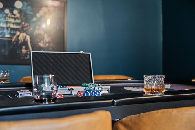 Casino-style poker table in a dim lounge with an open chip case, stacks of red, blue and green poker chips, playing cards and whiskey glasses on the felt