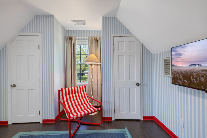 Cozy attic reading nook with blue-and-white striped wallpaper, red-trimmed dark hardwood floor, bold red-and-white striped lounge chair, floor lamp by a window overlooking a leafy yard, and a wall-mounted TV displaying a misty field.