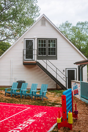 White gable-roof house exterior with black metal stairs to a second-floor door, four turquoise Adirondack chairs on the lawn, a large red outdoor play mat and oversized Connect Four game in a suburban backyard with trees