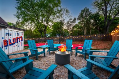 Cozy backyard at dusk with teal Adirondack chairs around a fire pit, red outdoor basketball court and string lights among trees