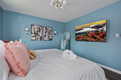 Cozy modern bedroom with teal-blue walls, crisp white bedding, frilly pink pillows and striped bolster, rolled towels on the bed, abstract wall art, arched floor mirror, brass sconces and a wall-mounted TV.