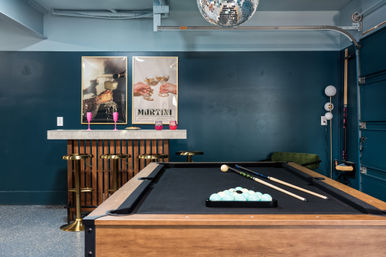 Stylish basement game room with wooden pool table and racked balls, teal painted walls, marble-top bar with gold stools and pink glasses, disco ball and framed martini posters.