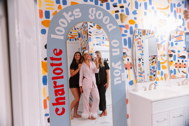 Three people in pajamas take a mirror selfie in a playful, colorful bathroom with abstract painted walls, an arched mirror with upbeat lettering, and a white double-sink vanity.