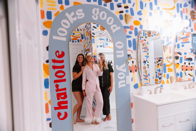 Three people in pajamas take a mirror selfie in a playful, colorful bathroom with abstract painted walls, an arched mirror with upbeat lettering, and a white double-sink vanity.
