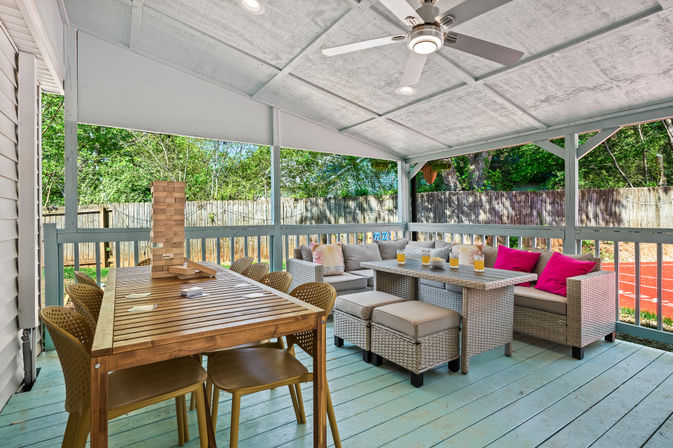 Sunlit covered backyard patio with a wooden dining table topped by a giant Jenga tower, rattan sectional with bright pink pillows and iced drinks, ceiling fan, light-blue painted deck and fenced yard with trees.
