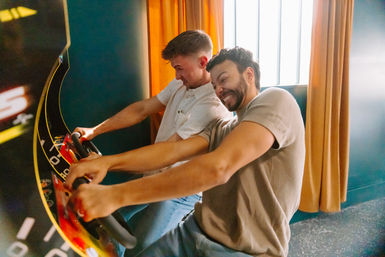 Two friends leaning in and laughing as they race on a two-player arcade driving game in a bright indoor gaming lounge with orange curtains.