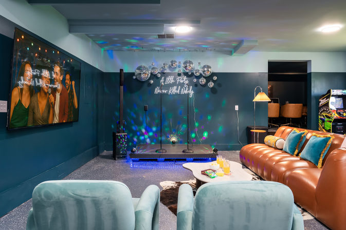 Vibrant basement karaoke lounge with a small raised stage, two microphone stands, cluster of disco balls and neon sign reading “A Little Party Never Killed Nobody,” teal walls, tufted leather sofa with teal pillows, velvet armchairs, large wall TV showing people singing, arcade machine and coffee table with drinks.