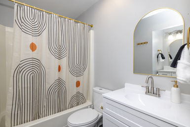 Bright contemporary bathroom with white vanity and sink, arched gold-framed mirror, brushed nickel faucet, toilet, and a playful geometric shower curtain featuring black arch lines and orange dots.
