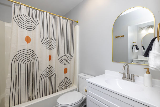Bright contemporary bathroom with white vanity and sink, arched gold-framed mirror, brushed nickel faucet, toilet, and a playful geometric shower curtain featuring black arch lines and orange dots.