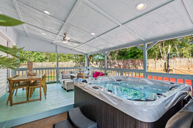 Covered backyard deck with a large hot tub, wicker lounge seating with colorful pillows, wooden dining table, ceiling fan, and fenced yard with basketball hoop