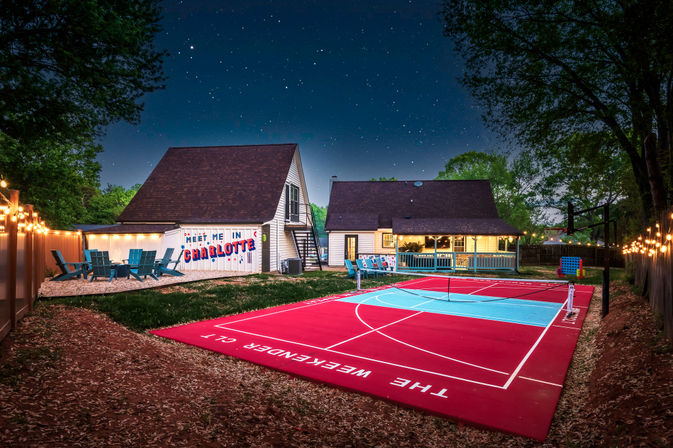 Starry-night backyard in Charlotte with two gabled cottages, a bright red-and-teal outdoor pickleball/tennis court, string lights, Adirondack seating and a painted 'Meet Me in Charlotte' mural.