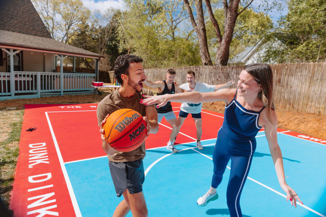 Four friends playing basketball on a sunny backyard half-court: a man clutching an orange ball while a smiling woman in blue athletic wear playfully reaches to block him, two teammates cheering near a wooden fence and porch.