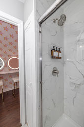 White marble-look tiled shower with glass sliding door, stainless rain showerhead and three amber pump bottles; view into adjacent pink star-patterned vanity with round lighted mirrors and stools.