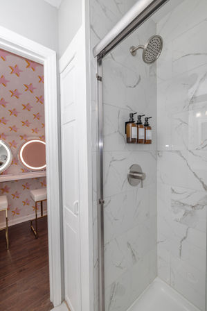 White marble-look tiled shower with glass sliding door, stainless rain showerhead and three amber pump bottles; view into adjacent pink star-patterned vanity with round lighted mirrors and stools.