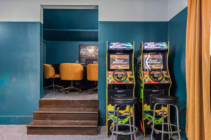 Vibrant game room with teal walls featuring two racing arcade cabinets with steering wheels, black bar stools, wooden steps leading to a raised lounge with mustard swivel chairs, and a mustard curtain at right.